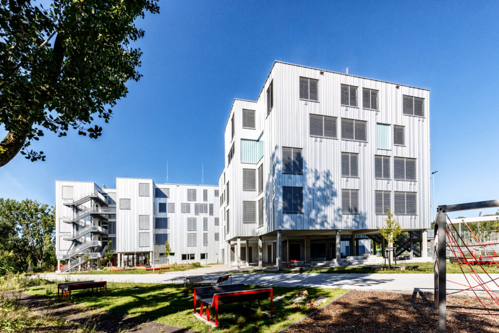 Mehrere moderne, weiße Bürogebäude mit großen Fenstern und umlaufenden Balkonen, umgeben von Bäumen und einer Grünfläche mit Sitzbänken und Spielgeräten.