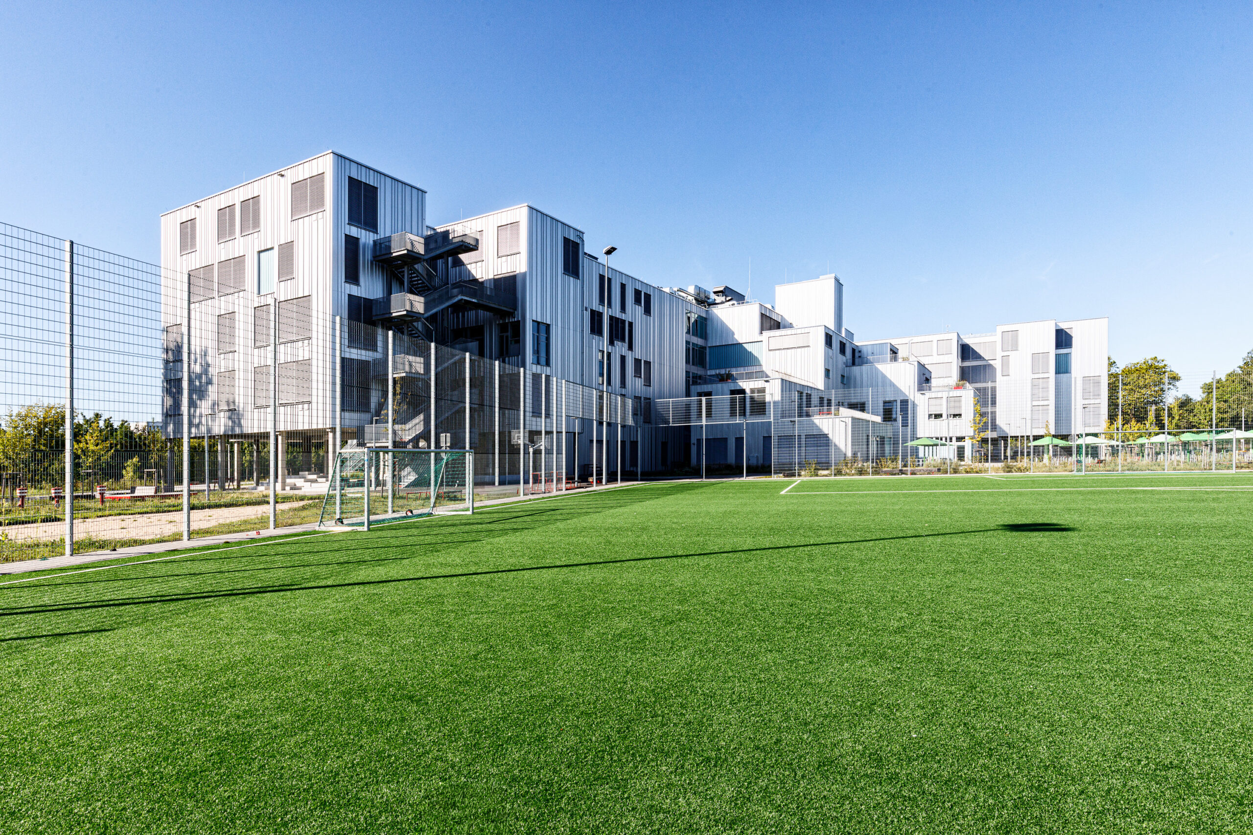 Moderner mehrstöckiger Schulgebäudekomplex mit großen Fenstern und Außentreppen, davor ein grünes Fußballfeld mit Toren und Zaun.