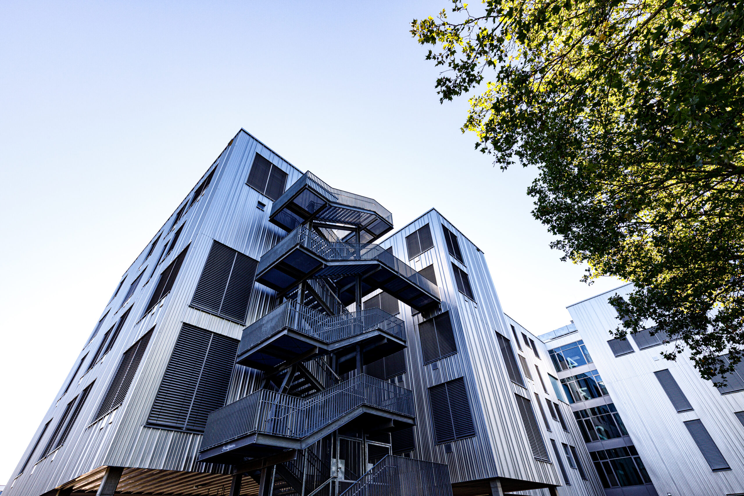 Blick von unten auf ein modernes, mehrstöckiges Gebäude mit außenliegender, metallener Treppe und Fenstern, umgeben von Bäumen.