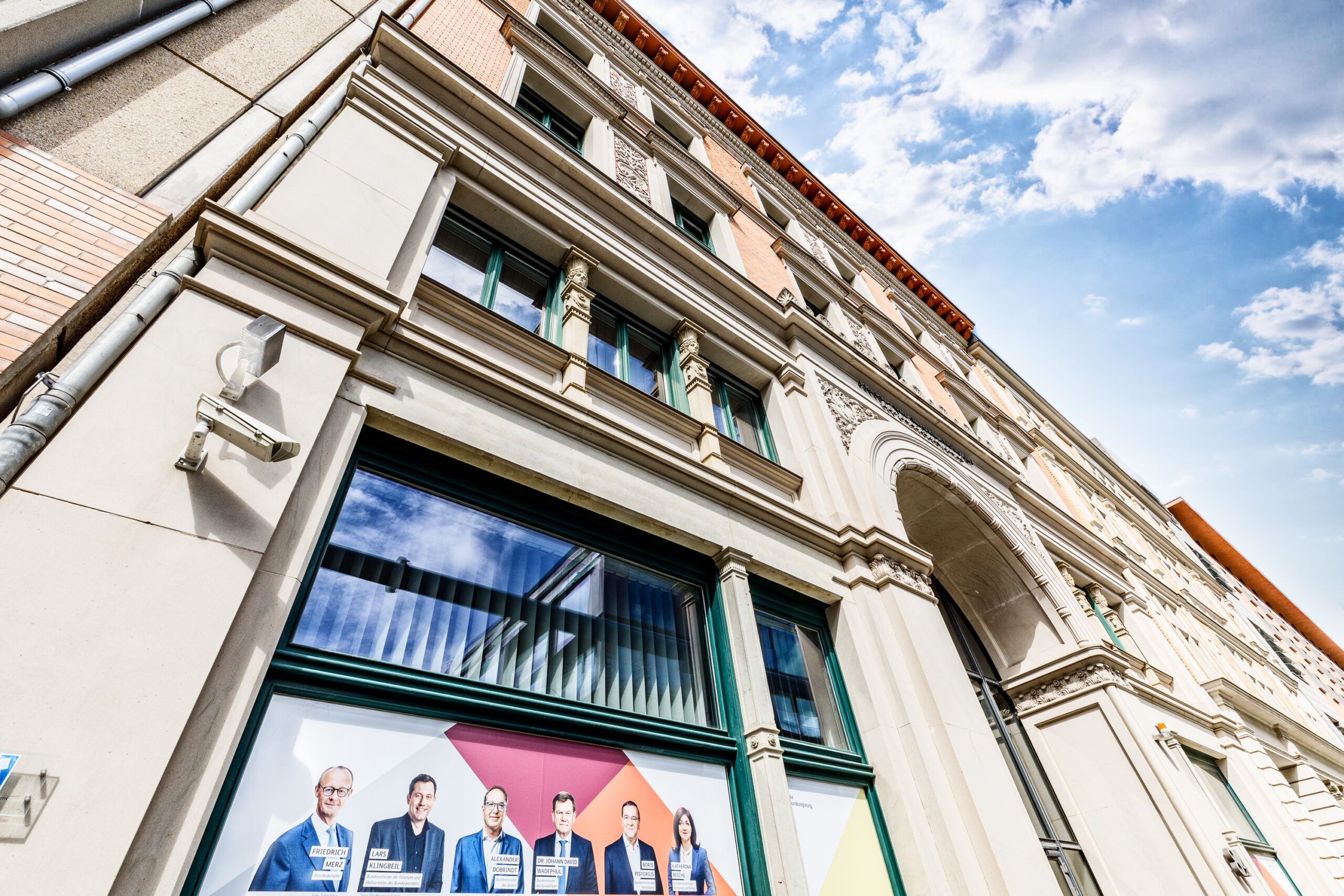 Fassade eines historischen Gebäudes mit großen Fenstern, an denen ein Werbebanner mit fünf Personen in Anzügen angebracht ist. Sicherheitskamera an der Wand sichtbar.