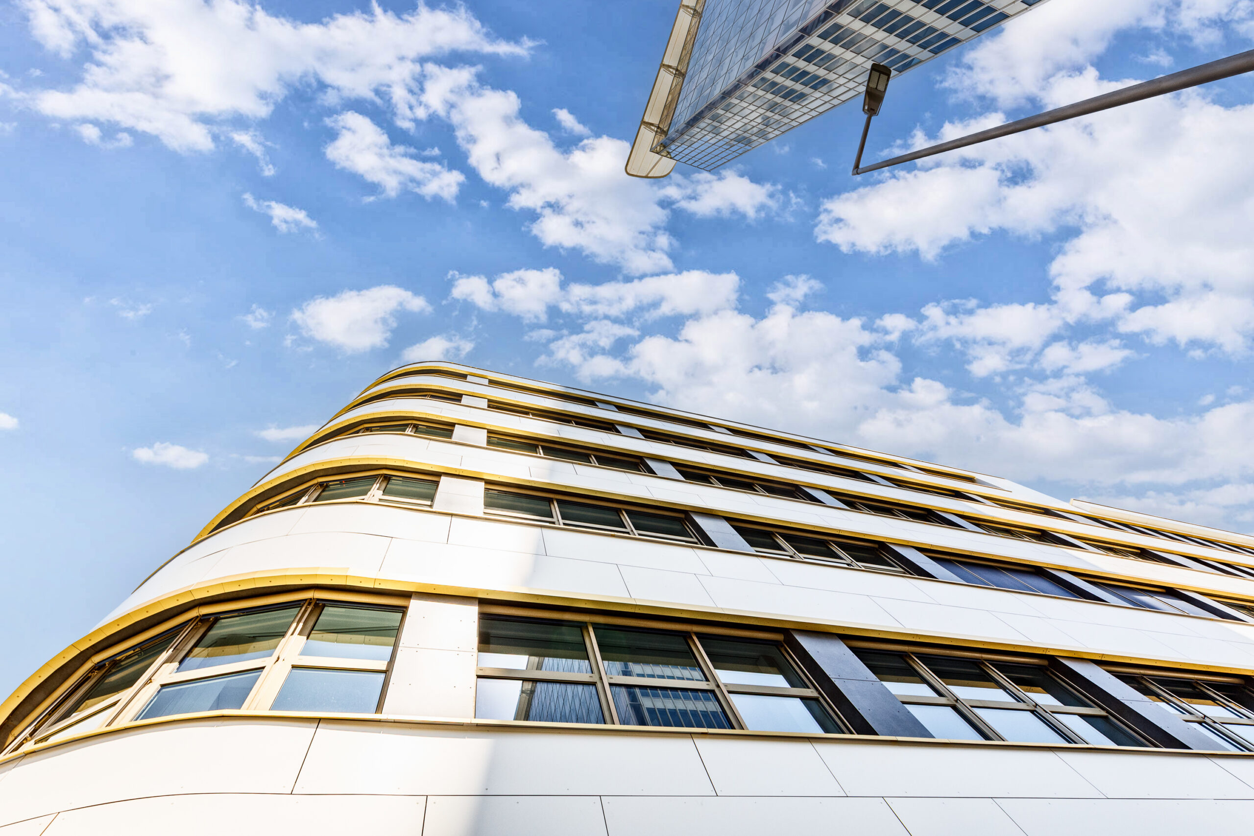 Moderne Architektur eines Gebäudes mit geschwungener Fassade, großen Fenstern und goldenen Akzenten vor blauem Himmel mit Wolken.