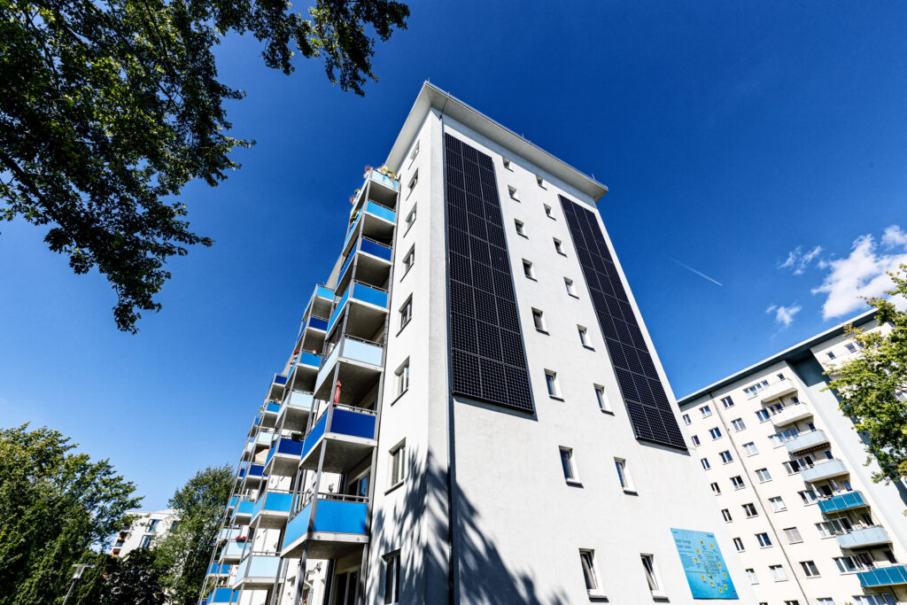 Modernes Wohngebäude mit blauen Balkonen und Photovoltaikanlage an der Fassade, vor strahlend blauem Himmel.