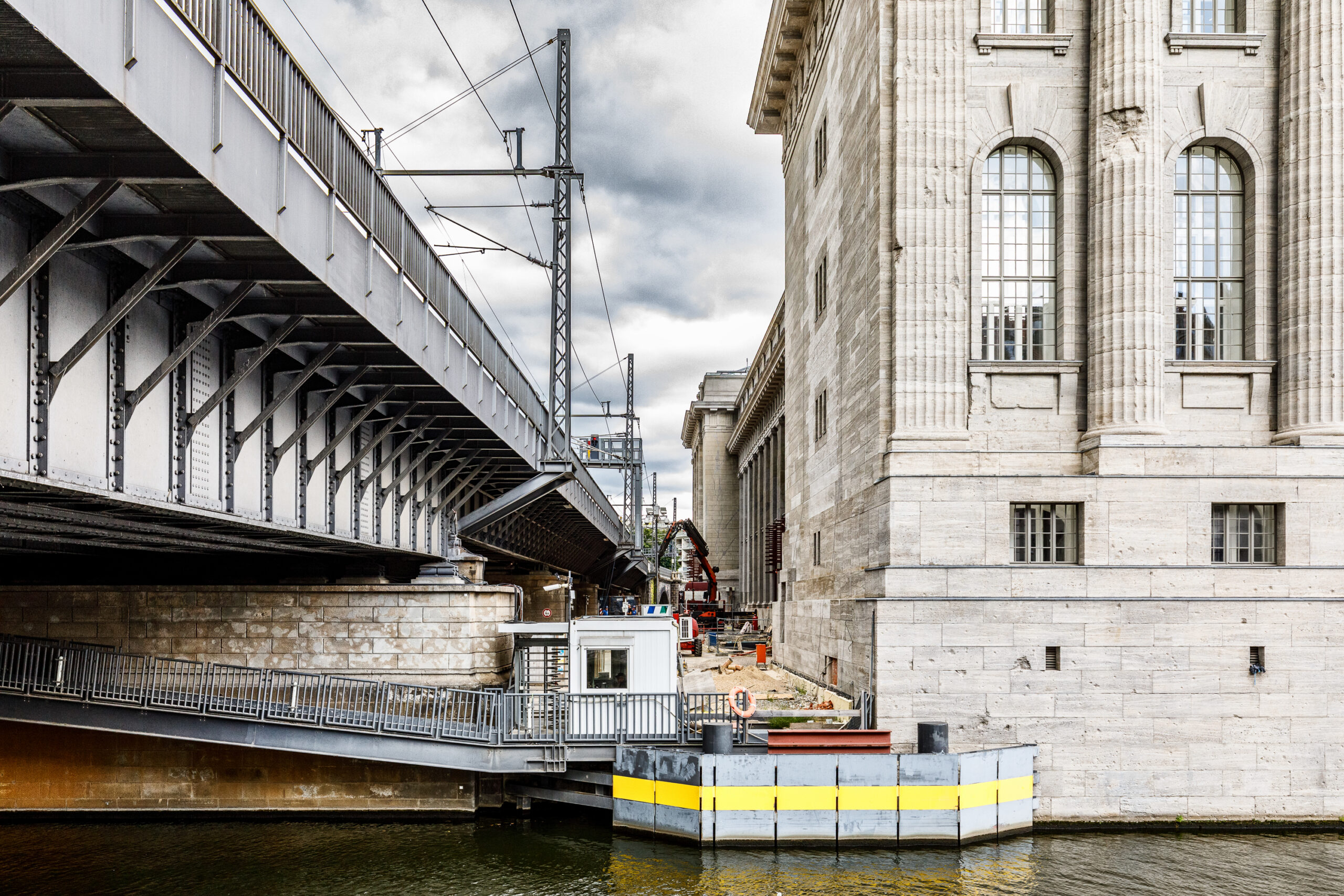 Blick auf eine Stahlbrücke über einen Fluss, neben einem historischen Gebäude mit Säulen. Im Vordergrund ist ein schwimmendes Dock mit einem kleinen Baucontainer zu sehen.