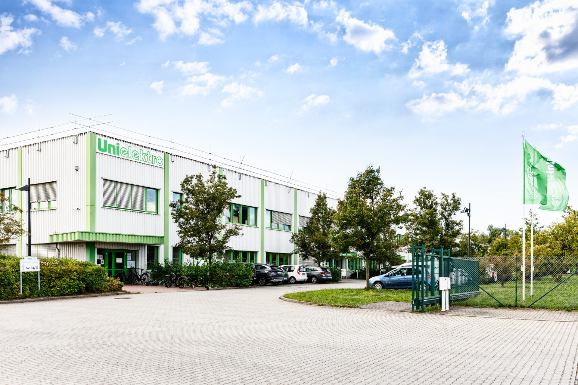 Fassade des UniElektro-Gebäudes mit grünen Akzenten, umgeben von Bäumen und einem Parkplatz.
